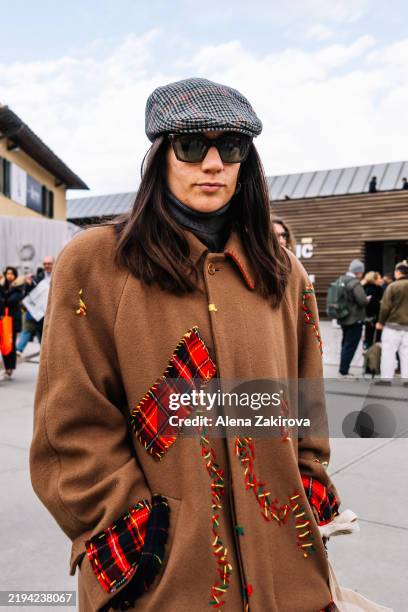 Guest wearing a brown coat with red plaid patches, a gray newsboy cap, sunglasses, blue jeans, and a white tote bag is seen during Pitti Immagine...