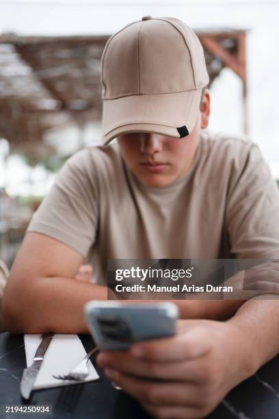 young man focused on his mobile phone while sitting at a café - boy cap stock pictures, royalty-free photos & images