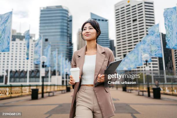 joven empresaria asiática bebiendo café para llevar mientras sostiene una computadora portátil en darling harbor en sydney en australia - cultura australiana fotografías e imágenes de stock