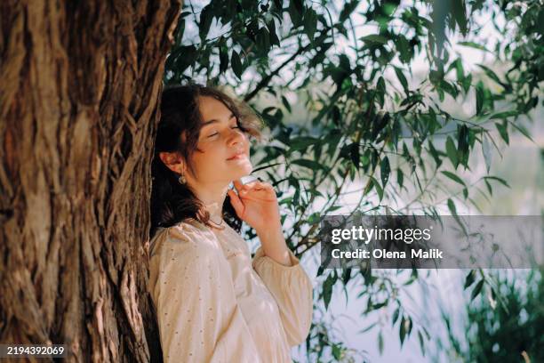young woman enjoying nature amid sunlight and shadows under a tree - frau kleid stock-fotos und bilder