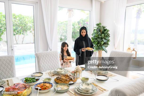 emiratische mutter und tochter decken tisch für familienessen, halten teller - speisezimmer stock-fotos und bilder