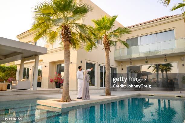 emirati man pointing towards large modern building with wife - villa stock pictures, royalty-free photos & images