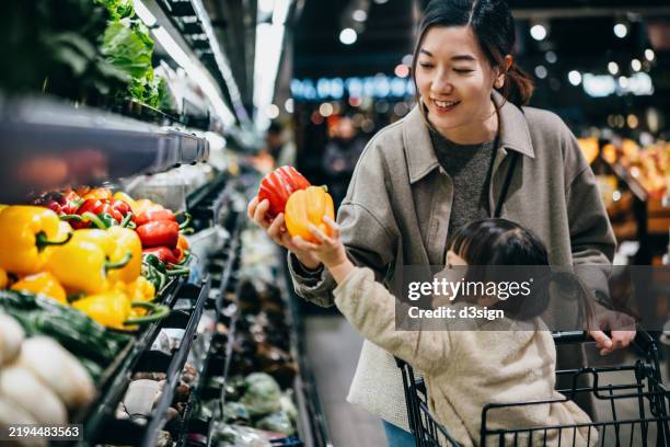 asian mother and daughter shopping for fresh fruits and vegetables together joyfully in supermarket. they are picking up some fresh bell peppers along the produce aisle. healthy eating lifestyle and habits. making a healthier food choice for family - lebensmittel einkauf stock-fotos und bilder
