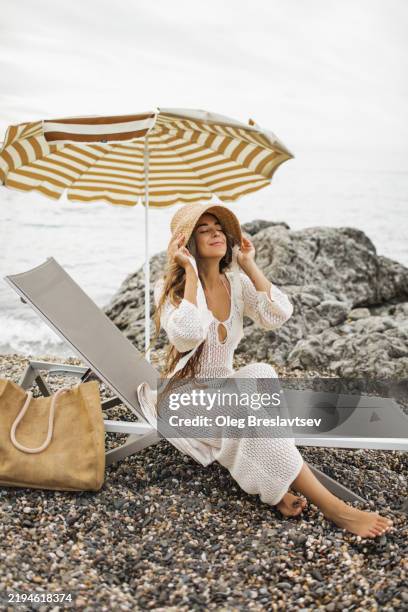 happy woman in straw hat relaxing on a scenic pebbled beach under a striped beach umbrella - túnica fotografías e imágenes de stock