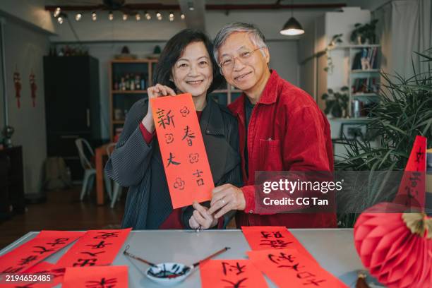 portrait senior couple holding chinese couplet practising chinese calligraphy for chinese new year fai chun (auspicious messages) looking at camera smiling - ink and brush stock pictures, royalty-free photos & images