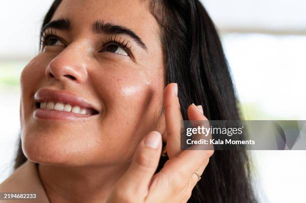 smiling woman applying moisturizer on face - liscio foto e immagini stock