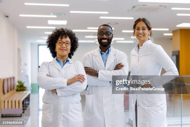 multi-ethnic medical team smiling with arms crossed in hospital corridor - lab coat stock pictures, royalty-free photos & images
