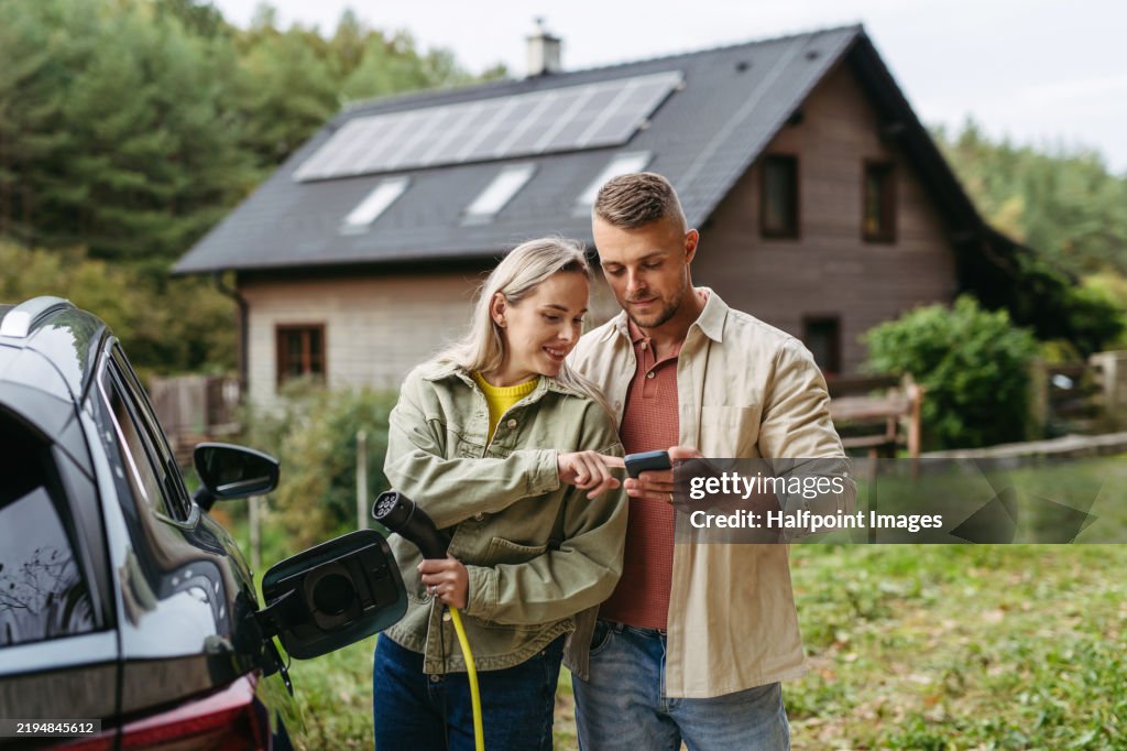 Couple charging their electric car in front of house. Checking charging progress on smartphone.