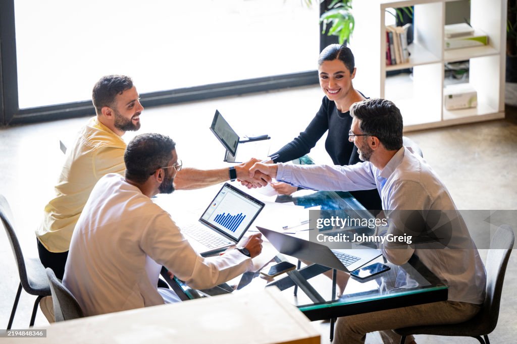 Vista desde alto de dos hombres de negocios dándose la mano a través de la mesa, dos colegas mirando y sonriendo