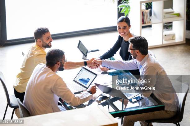 high angle view of two businessmen shaking hands across table, two colleagues watching and smiling - woman-man-handshake-across-table stock pictures, royalty-free photos & images