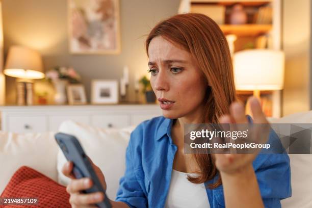 confused woman looking at smartphone sitting on sofa at home - redhead stock pictures, royalty-free photos & images
