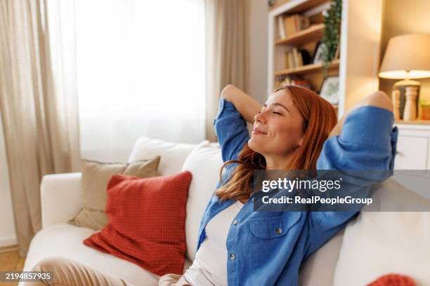 junge frau, die sich auf dem sofa mit den händen hinter dem kopf entspannt - sofa stock-fotos und bilder