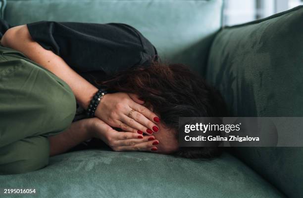 depressed brunette young woman face covered with hands sitting on sofa at home - menstruation stock pictures, royalty-free photos & images
