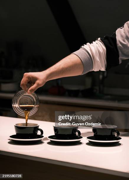barista pouring freshly brewed filter coffee into three black ceramic cups, modern café setting with warm lighting, specialty coffee preparation and artisan brewing techniques, coffee tasting experience - espresso stockfoto's en -beelden