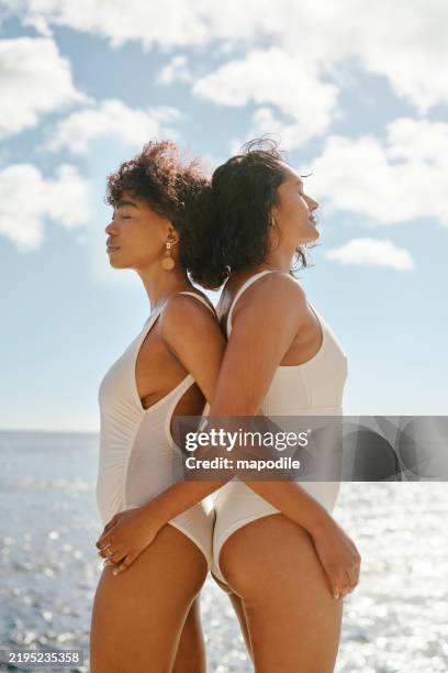 vista lateral de dos mujeres elegantes en trajes de baño blancos en una playa soleada con el océano y el cielo en el fondo - baño-de-pies fotografías e imágenes de stock