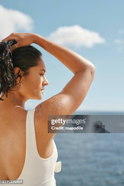 side portrait of woman in white swimwear with hands in her hair against a blue sky and ocean background - mid adult women stock pictures, royalty-free photos & images
