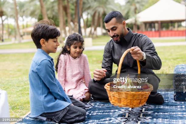happy middle eastern father placing fruit basket in front of kids at park picnic - wicker stock pictures, royalty-free photos & images