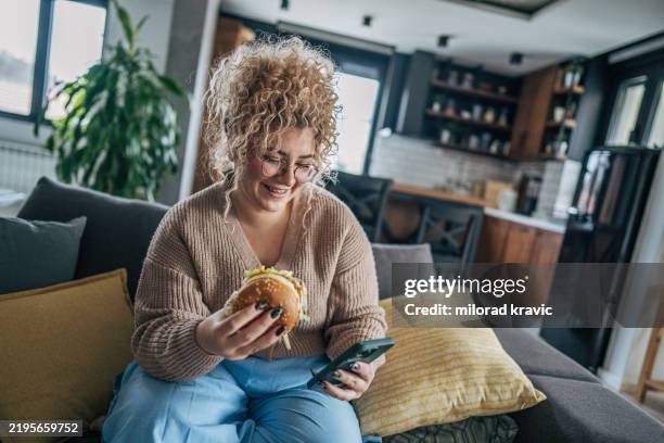 a woman with curly hair holds a burger in one hand and a phone in the other - messy cheeseburger stock pictures, royalty-free photos & images