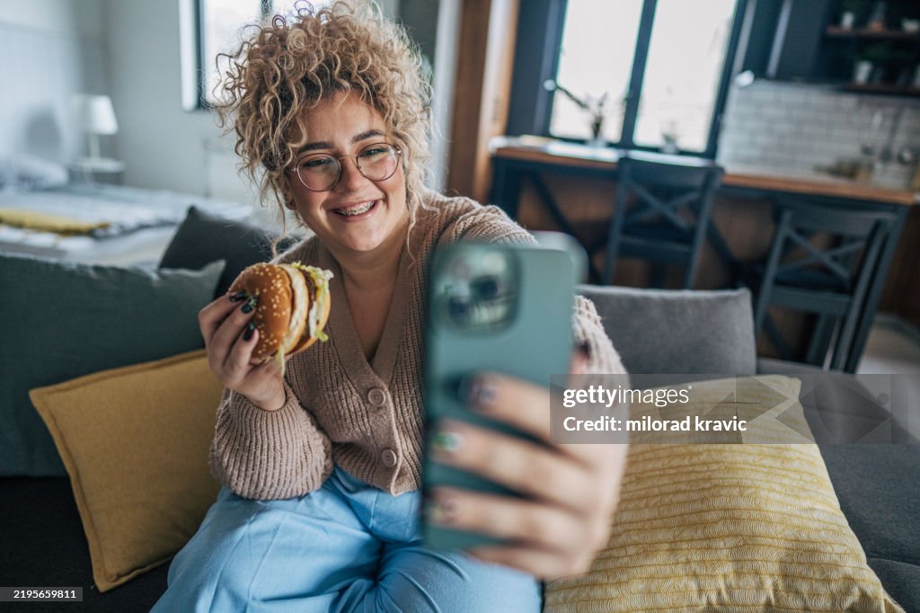 Happy woman enjoying a hamburger on the couch while taking a selfie