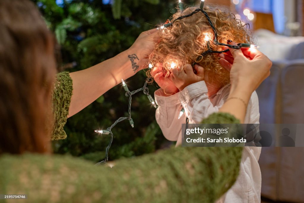 Mother playfully places a string of Christmas lights on a child's head in a cozy indoor setting, Ohio, USA