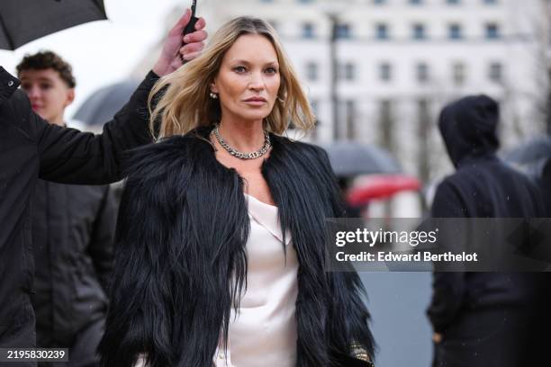 Kate Moss wears silver earrings, silver choker necklace, black faux fur coat, mixed metal layered bangle bracelets, a gold ring, outside Dior Homme,...