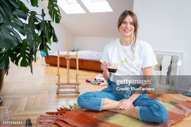 young woman taking a coffee break while painting in her home studio - de-pernas-cruzadas imagens e fotografias de stock