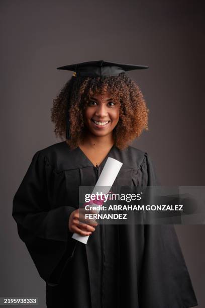 young graduate wearing a gown and mortarboard, proudly holding her diploma and smiling brightly at the camera against a gray background - female likeness stock pictures, royalty-free photos & images
