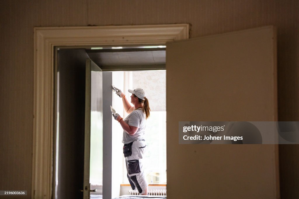 Female plasterer applying putty on wall seen through doorway