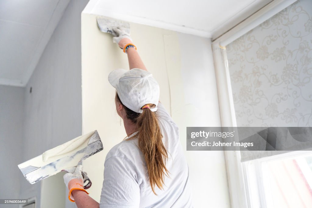 Rear view of female painter applying putty on wall at workplace