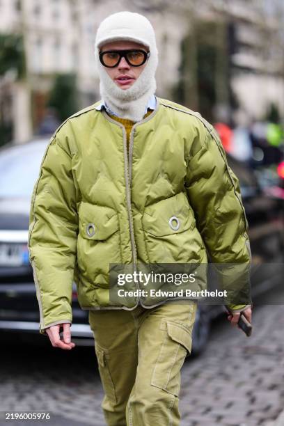 Guest wears yellow and black sunglasses, a white wool balaclava, a light green oversized jacket, green pants, outside Hermes, during the Menswear...