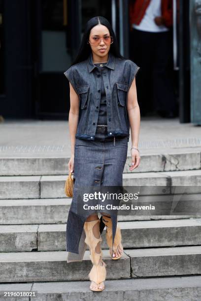 Aleali May wears blue denim Courreges top, matching vest and midi skirt, beige fur sandal boots, beige woven bag, outside Sacai, during the Menswear...