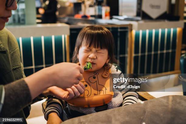 displeased little asian toddler girl refusing to eat vegetables while her mother is feeding her at dining table. children growth and development. kids health, healthy eating lifestyle and nutrition concepts - grimace de crispation photos et images de collection