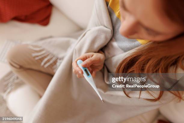 sick young woman checking her temperature while sitting on the sofa - koorts stockfoto's en -beelden