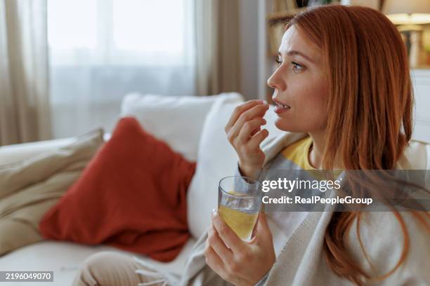 joven enferma tomando una pastilla y bebiendo un vaso de agua en el sofá - tomar medicina fotografías e imágenes de stock