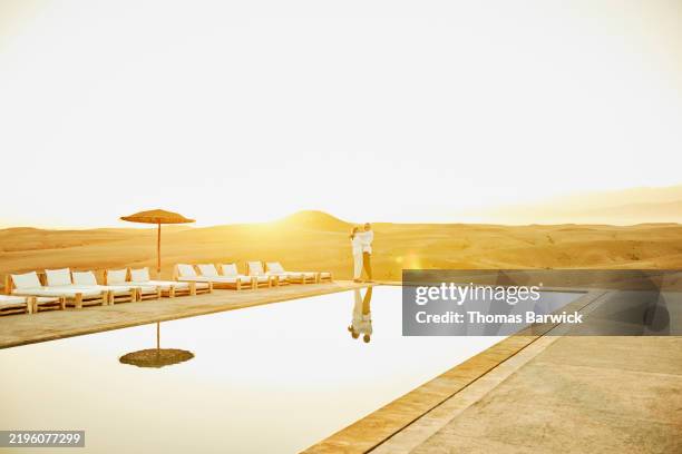 extreme wide shot senior couple embracing poolside at desert resort - mittlere entfernung stock-fotos und bilder