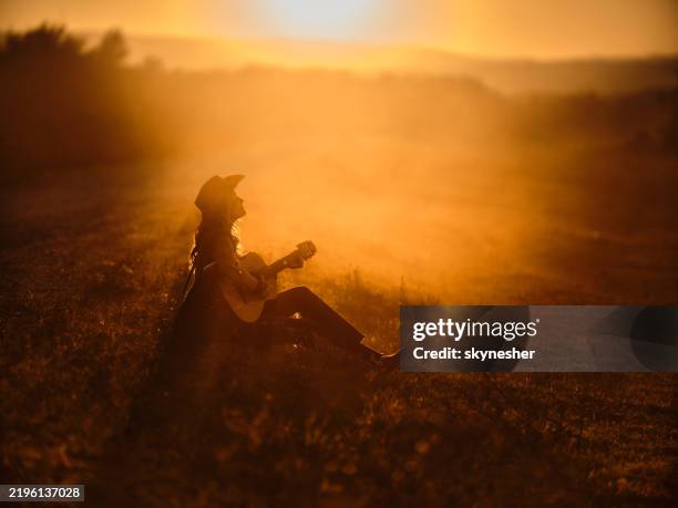 relaxed cowgirl playing a guitar in a meadow at sunset. - gitarist stockfoto's en -beelden