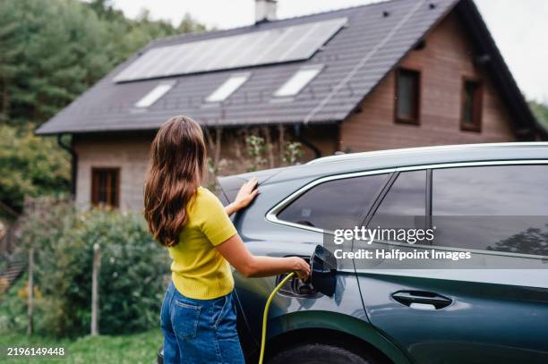 young woman is charging her electric car, parked in front of a house with solar panels on the roof. - battery life stock pictures, royalty-free photos & images