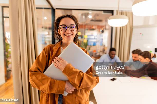 jeune femme d’affaires tenant un ordinateur portable souriant à la caméra dans un bureau moderne - jeune adulte photos et images de collection