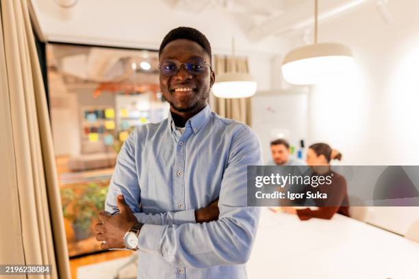 selbstbewusster junger geschäftsmann, lächelnd mit verschränkten armen in modernem büro - halbnahe-einstellung stock-fotos und bilder