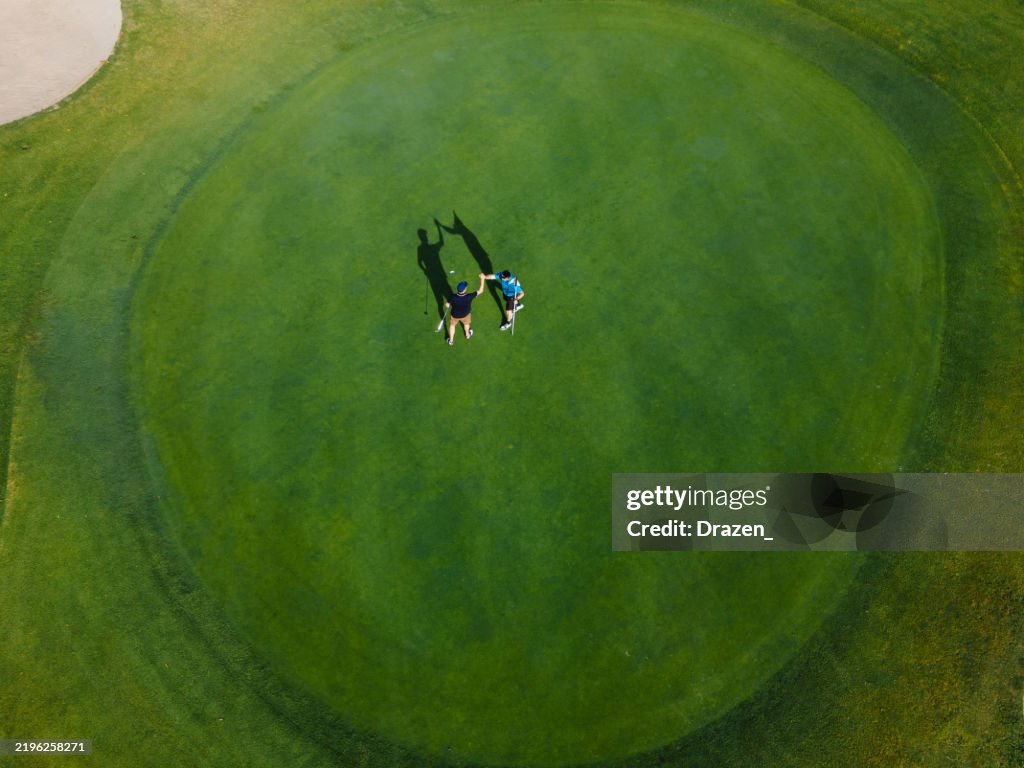 Zwei Golfer spielen auf einem leuchtend grünen Golfplatz mit Sandbunker und umliegenden Bäumen unter klarem Himmel