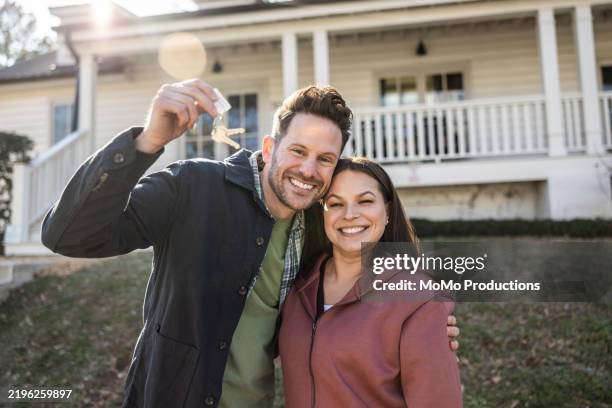 portrait of young couple holding house keys in front of their new home - proprietario di casa foto e immagini stock