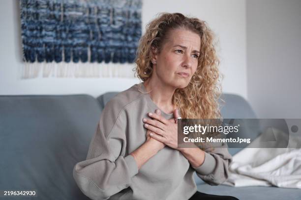 anxious middle-aged woman holging hands on chest feeling stressed, mature woman sitting on sofa at home suffering from panic attack - menselijk hart stockfoto's en -beelden