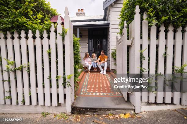 family sitting on porch of charming house with white fence - tuinhek stockfoto's en -beelden