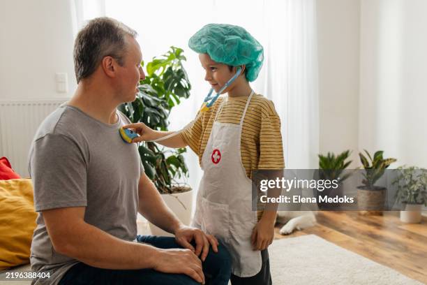 a father and son play doctor together, laughing and enjoying their time. the child uses a toy stethoscope to check his dad - vestita da dottoressa foto e immagini stock