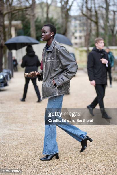 Model wears dark gray oversized leather jacket, light blue flared denim jean pants, shiny black pointed toe heeled boots leather shoes, outside...