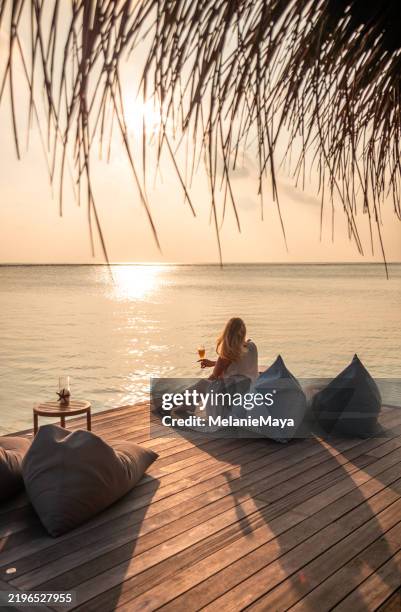 frau genießt sundowner trinken cocktail in der overwater lounge bar mit liegestuhl sitzsäcken und warmer goldener stunde sonnenuntergang licht in der idyllischen lagune des inselresorts der malediven - malediven stock-fotos und bilder
