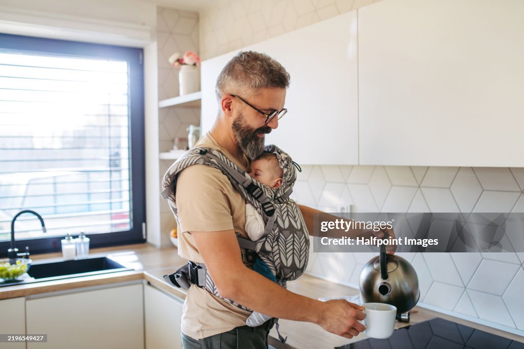 Mature father making a tea while newborn child sleeping in baby carrier. Concept of paternity leave and dad as caregiver for baby.
