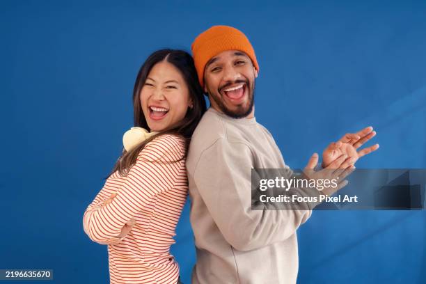 diverse young couple standing back-to-back, smiling candidly in studio setting, sharing playful moment against blue backdrop - back to back stock pictures, royalty-free photos & images