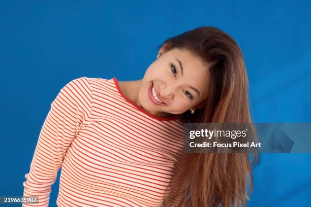 smiling young woman tilting head, long hair flowing, standing against bright blue backdrop with warm, friendly expression - head cocked stock pictures, royalty-free photos & images