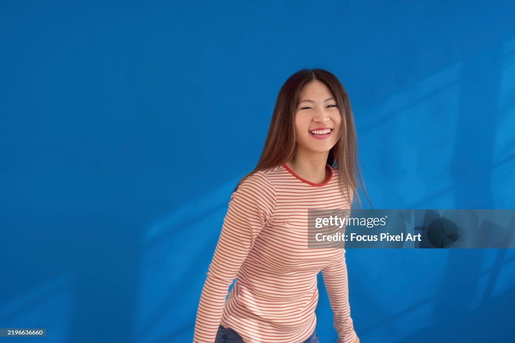 Smiling brunette leaning forward, wearing striped top, standing against bright blue background, radiating positive energy